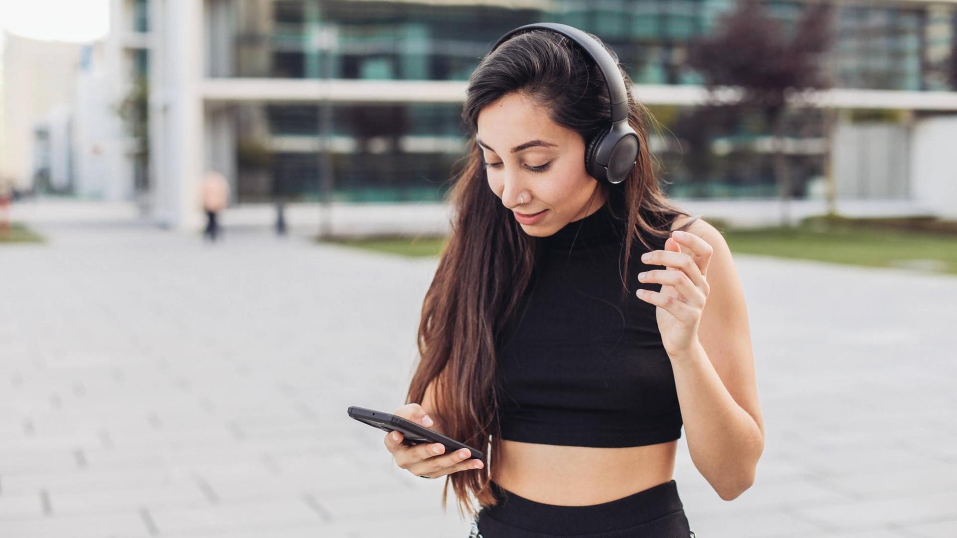 Junge Frau mit Kopfhörern schaut auf ihr Smartphone Junge Frau mit Kopfhörern schaut auf ihr Smartphone