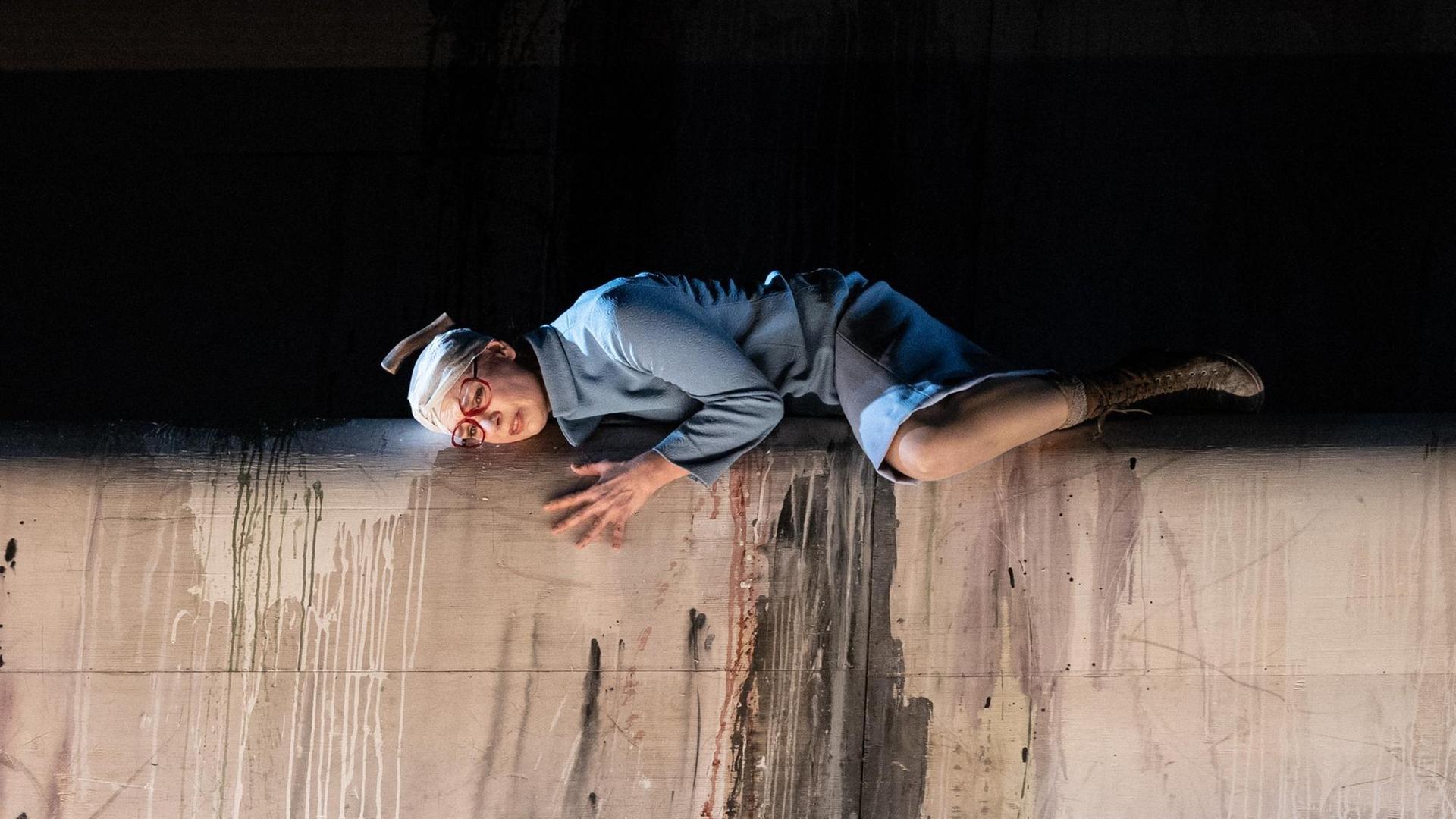 Szenenbild aus der Oper "Toteis" von Manuela Kerer & Matthias Plattner mit Isabel Seebacher als "Viktoria", die bäuchlings vor einem schwarzen Hintergrund auf einer grauen Mauer liegt und einen Verband um den Kopf trägt.