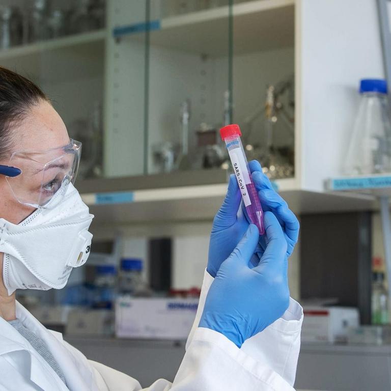 Eine Wissenschaftlerin mit Mundschutz, Schutzbrille und Handschuhen hält ein Laborröhrchen mit der Aufschrift "SARS-CoV 2" in den Händen. Eine Wissenschaftlerin mit Mundschutz, Schutzbrille und Handschuhen hält ein Laborröhrchen mit der Aufschrift "SARS-CoV 2" in den Händen.