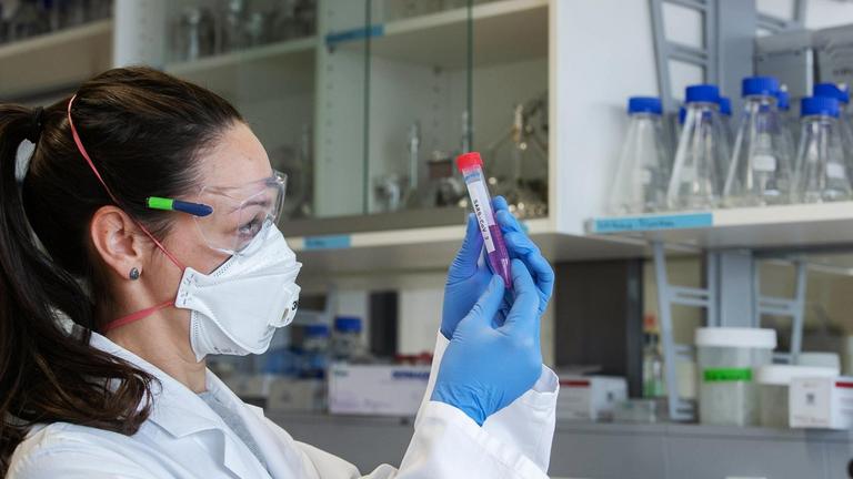 Eine Wissenschaftlerin mit Mundschutz, Schutzbrille und Handschuhen hält ein Laborröhrchen mit der Aufschrift "SARS-CoV 2" in den Händen.
