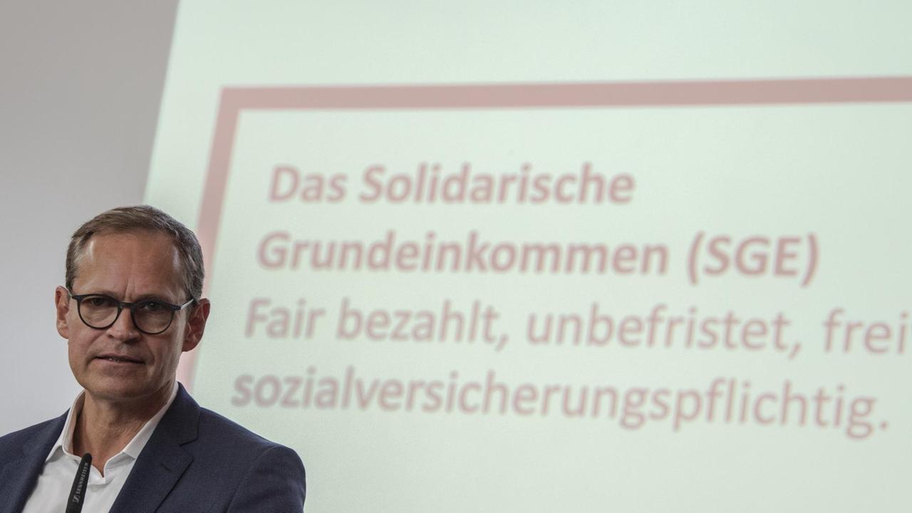 Der Regierende Bürgermeister Michael Müller (SPD) vor einer Wand, auf die mit einem Beamer die Schrift projiziert ist "Das Solidarische Grundeinkommen (SGE): fair bezahlt, frei[willig], sozialversicherungspflichtig"