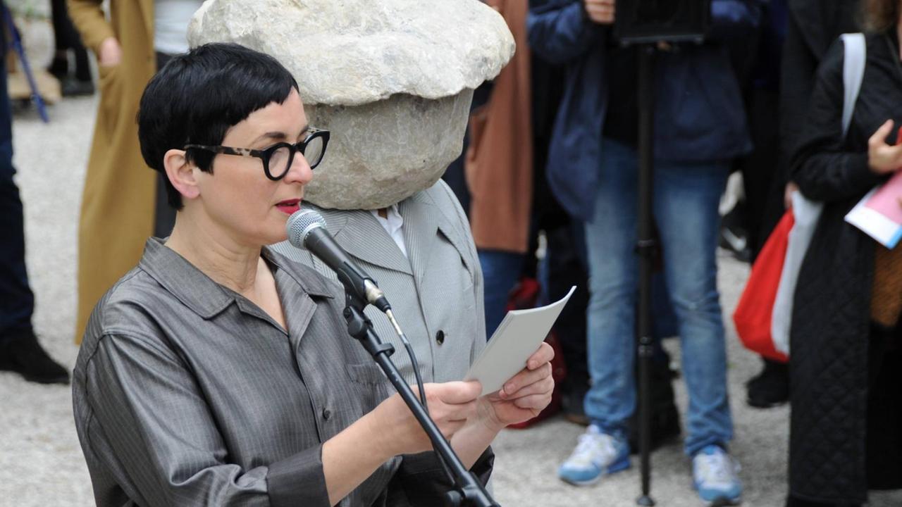 Helene Duldung verliest etwas von einem Stück Papier, während die Künstlerin Natscha Süder Happelmann, die eine Steinmaske über ihrem Kopf trägt, neben ihr steht. Helene Duldung verliest etwas von einem Stück Papier, während die Künstlerin Natscha Süder Happelmann, die eine Steinmaske über ihrem Kopf trägt, neben ihr steht.