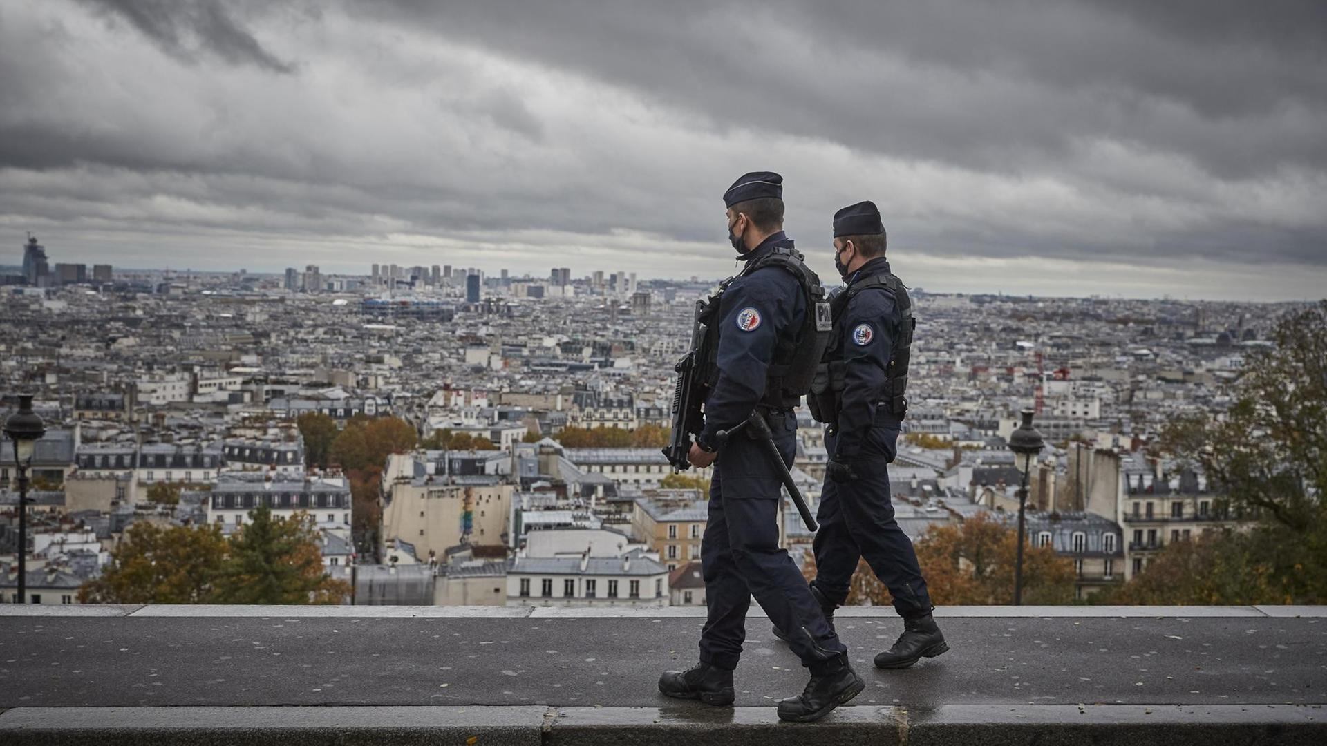 Bewaffnete Polizisten mit Masken laufen Patrouille auf dem Vorplatz der Pariser Basilika Sacré Cœur, im Hintergrund eine Aussicht auf die Dächer von Paris. Bewaffnete Polizisten mit Masken laufen Patrouille auf dem Vorplatz der Pariser Basilika Sacré Cœur, im Hintergrund eine Aussicht auf die Dächer von Paris.