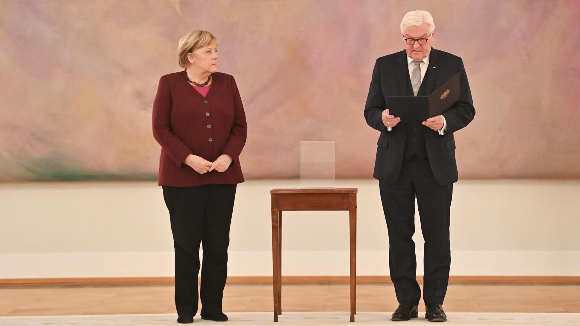 Bundespräsident Frank-Walter Steinmeier händigt Bundeskanzlerin Angela Merkel die Entlassungsurkunde aus.Beide stehen vor einem abstrakten rosa Gemälde, zwischen ihnen: ein hochbeiniger Beistelltisch. Bundespräsident Frank-Walter Steinmeier händigt Bundeskanzlerin Angela Merkel die Entlassungsurkunde aus.Beide stehen vor einem abstrakten rosa Gemälde, zwischen ihnen: ein hochbeiniger Beistelltisch.