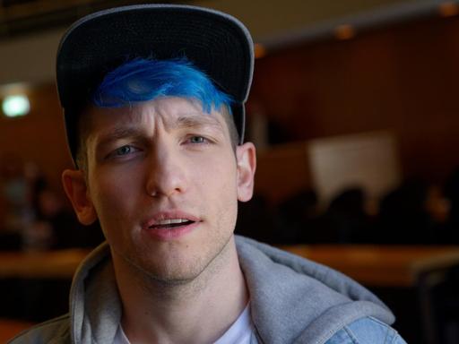 Youtuber Rezo, mit blau gefärbtem Haar und Basecap, schaut skeptisch in die Kamera, im Hintergrund: ein holzgetäfelter Veranstaltungssaal