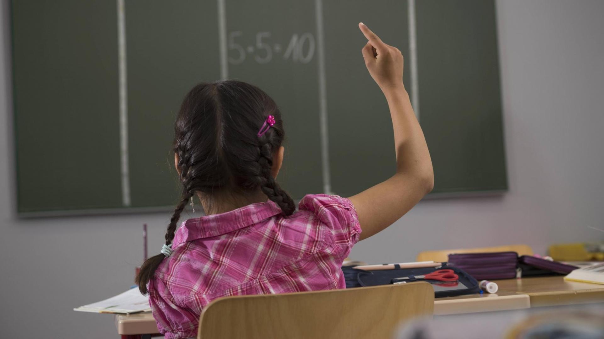 Ein von hinten fotografiertes Mädchen mit dunklen Zöpfen sitzt vor einer Tafel in einem Klassenraum und hebt seine rechte Hand. Ein von hinten fotografiertes Mädchen mit dunklen Zöpfen sitzt vor einer Tafel in einem Klassenraum und hebt seine rechte Hand.