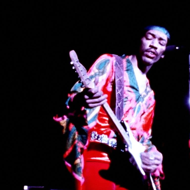 Jimi Hendrix steht mit Gitarre auf einer Bühne und trägt bunte Kleidung und ein Stirnband.