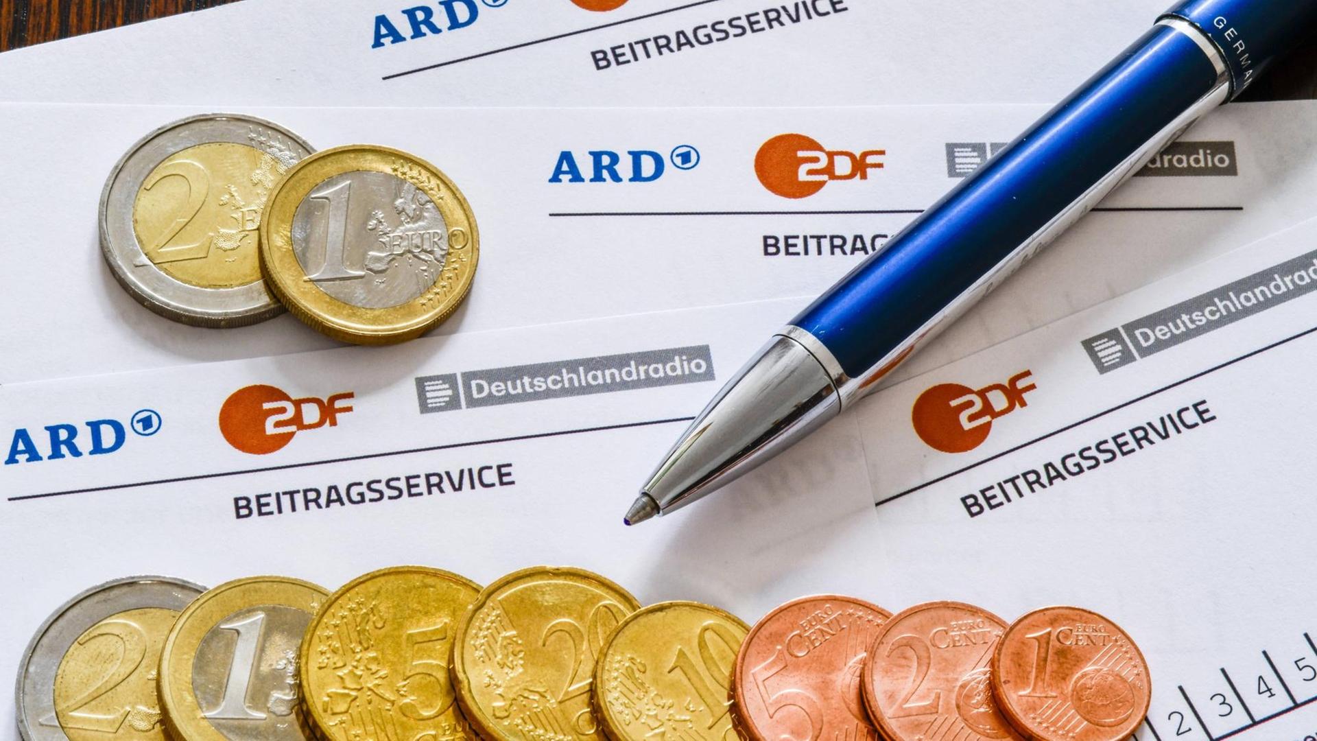 Auf mehreren Formularen des Beitragsservice für den Rundfunkbeitrag liegen Geldmünzen sowie ein Kugelschreiber. Auf mehreren Formularen des Beitragsservice für den Rundfunkbeitrag liegen Geldmünzen sowie ein Kugelschreiber.