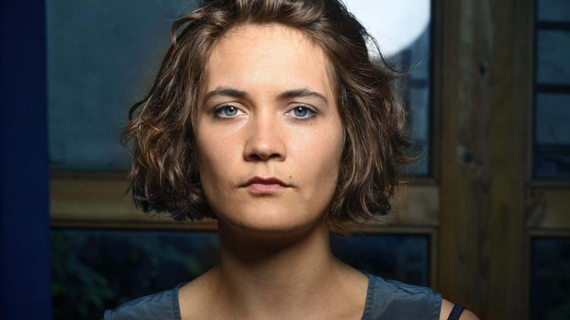Porträtansicht einer jungen Frau mit brünettem, halblangem Haar und blauen Augen. Sie schaut dem Betrachter ernst ins Gesicht. i Porträtansicht einer jungen Frau mit brünettem, halblangem Haar und blauen Augen. Sie schaut dem Betrachter ernst ins Gesicht. i