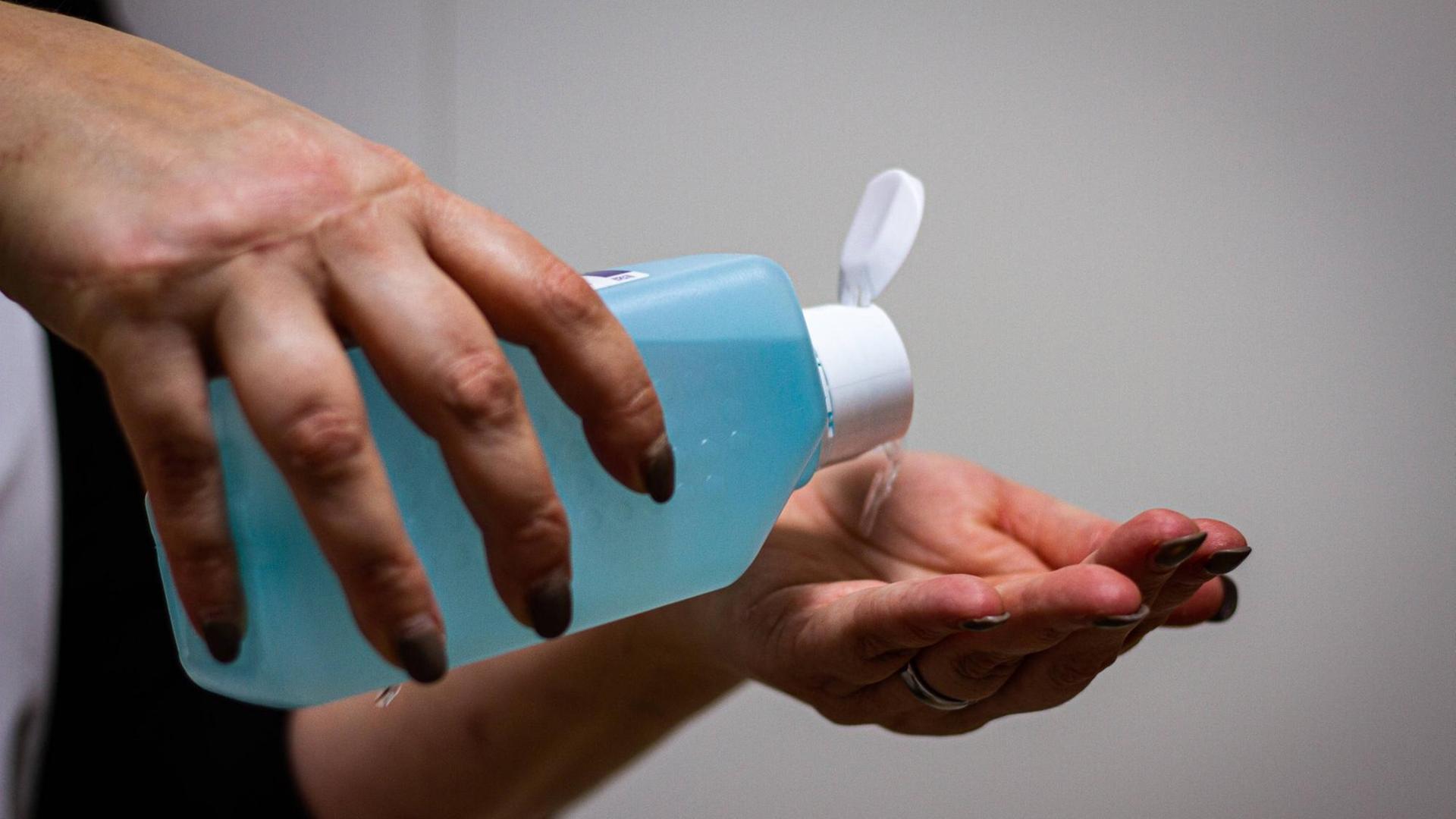 Eine Person hält eine große blaue Flasche Desinfektionsmittel in den Händen und kippt die Flüssigkeit in die linke Hand. Eine Person hält eine große blaue Flasche Desinfektionsmittel in den Händen und kippt die Flüssigkeit in die linke Hand.