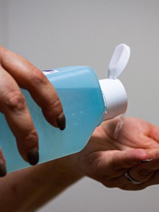 Eine Person hält eine große blaue Flasche Desinfektionsmittel in den Händen und kippt die Flüssigkeit in die linke Hand.