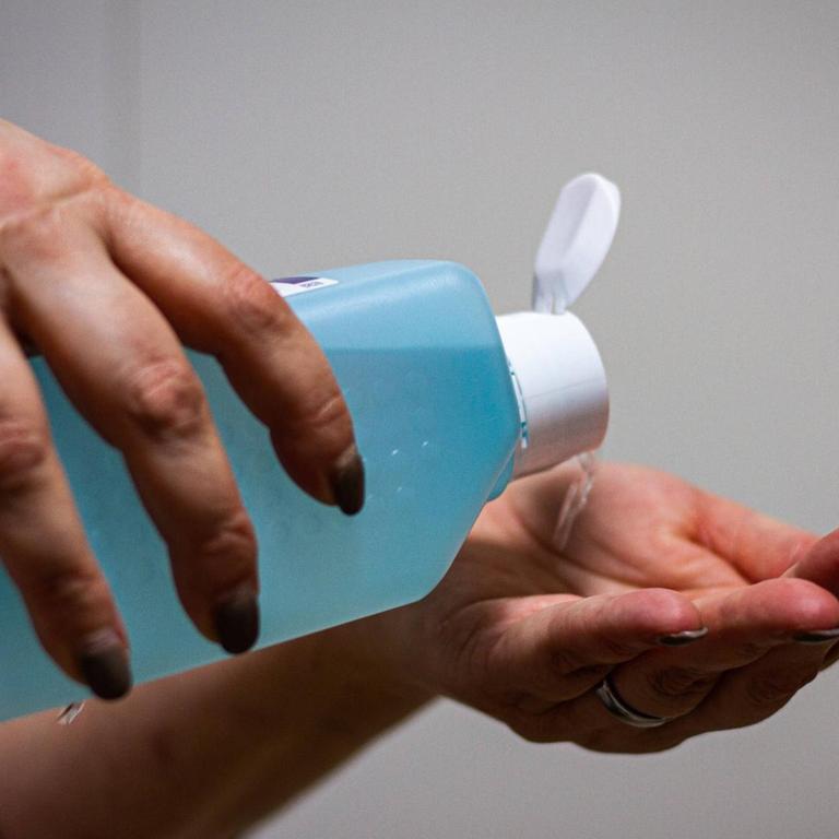 Eine Person hält eine große blaue Flasche Desinfektionsmittel in den Händen und kippt die Flüssigkeit in die linke Hand. 