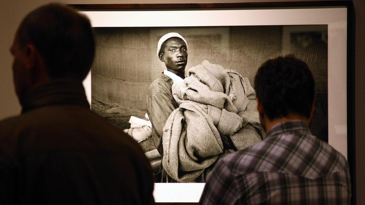 Zwei Menschen betrachten in einer Ausstellung in Berlin ein Bild des Fotografen Sebastião Salgado. Darauf zu sehen ist ein Mann mit Stoff in den Händen. Zwei Menschen betrachten in einer Ausstellung in Berlin ein Bild des Fotografen Sebastião Salgado. Darauf zu sehen ist ein Mann mit Stoff in den Händen.