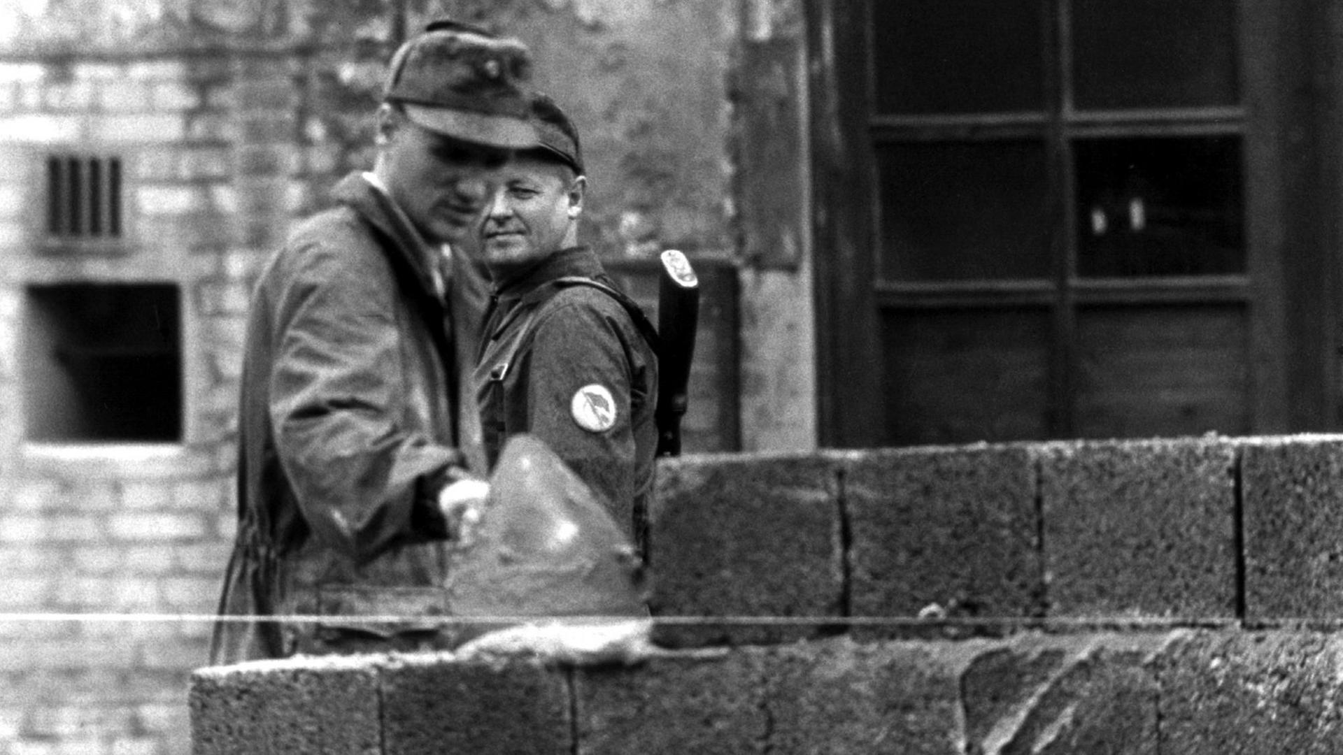Ein Bauarbeiter fügt 1961 Steine zur Berliner Mauer hinzu, ein Volkspolizist beobachtet ihn dabei. Ein Bauarbeiter fügt 1961 Steine zur Berliner Mauer hinzu, ein Volkspolizist beobachtet ihn dabei.