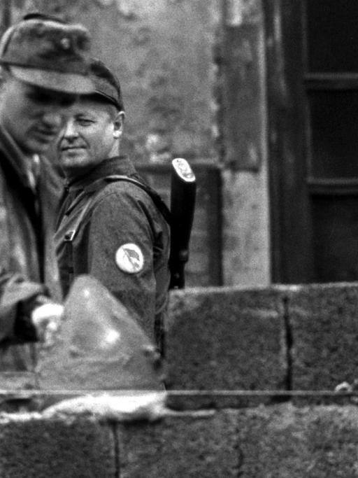 Ein Bauarbeiter fügt 1961 Steine zur Berliner Mauer hinzu, ein Volkspolizist beobachtet ihn dabei.
