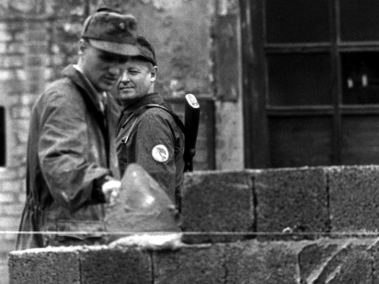 Ein Bauarbeiter fügt 1961 Steine zur Berliner Mauer hinzu, ein Volkspolizist beobachtet ihn dabei.