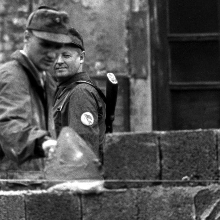 Ein Bauarbeiter fügt 1961 Steine zur Berliner Mauer hinzu, ein Volkspolizist beobachtet ihn dabei.