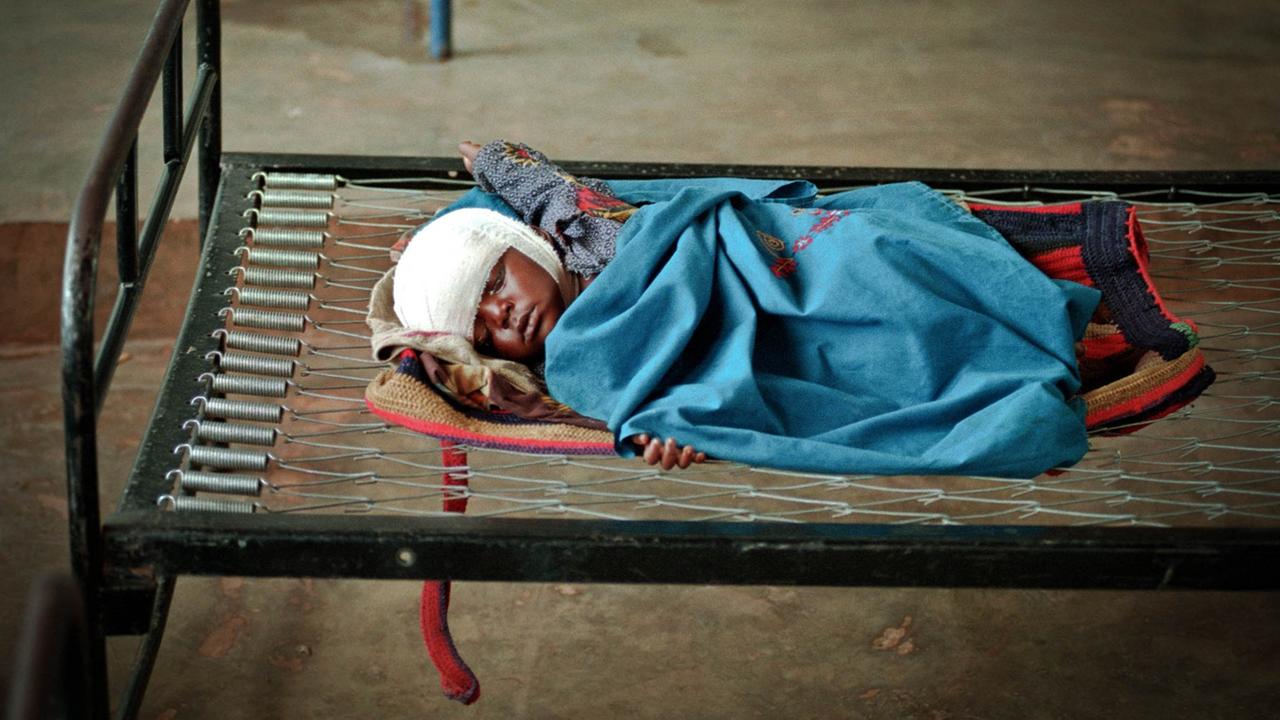 Ein Kleinkind liegt mit verbundenen Kopf auf einem eisernen Bettgestell in eine blaue Decke gehüllt und schläft.