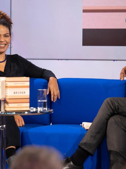 Jackie Thomae sitzt auf dem blauen Sofa mit René Aguigah, auf dem Tisch steht ihr Buch "Brüder".