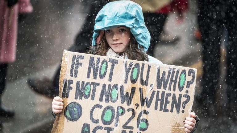 Ein junges Mädchen mit grüner Regenjacke trägt ein Pappschild, auf dem steht: "If not you, who? If not now, when? COP26 act now!! Es regnet. Ein junges Mädchen mit grüner Regenjacke trägt ein Pappschild, auf dem steht: "If not you, who? If not now, when? COP26 act now!! Es regnet.