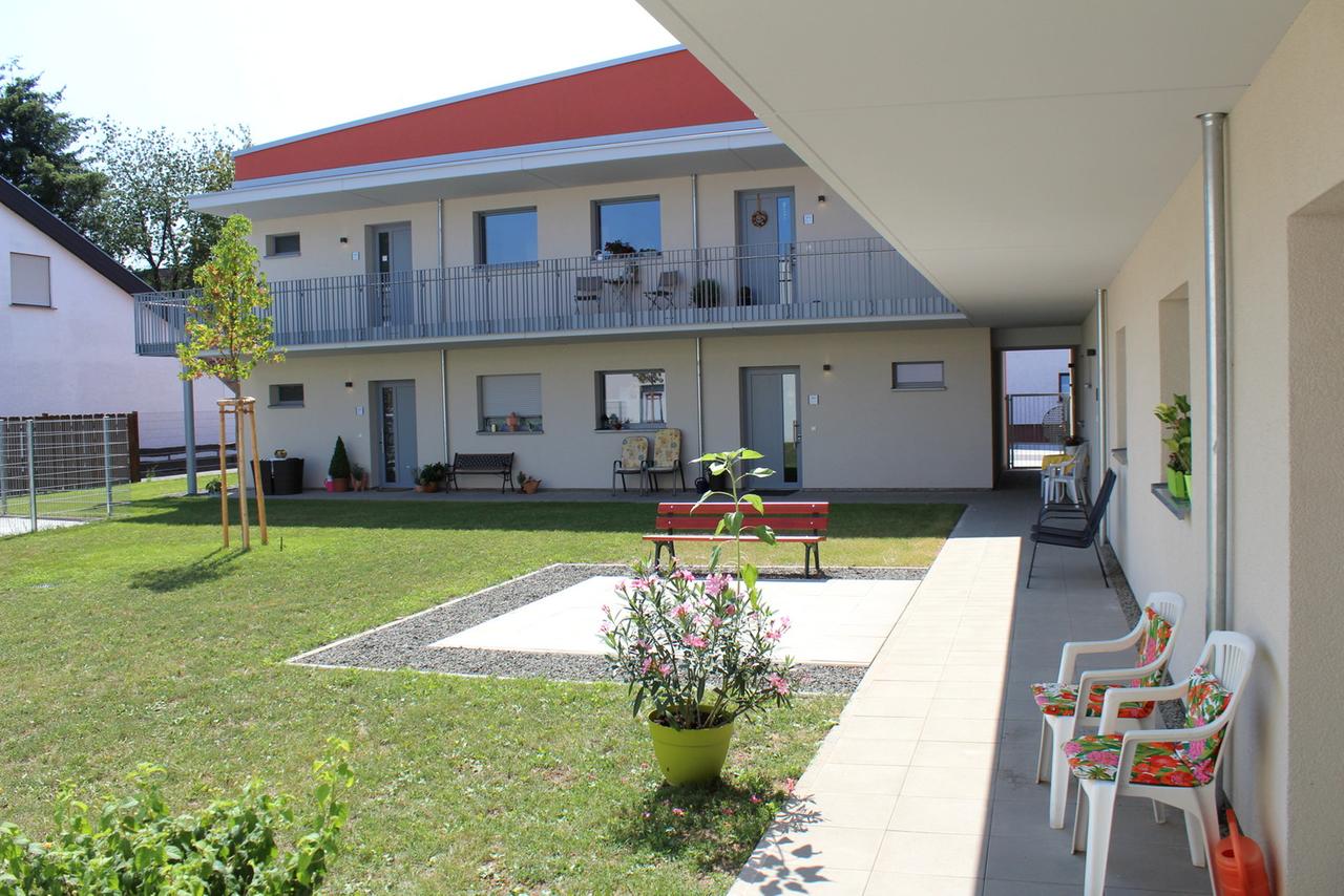 Das Seniorendorf vom Innenhof aus. Neben einer Grünfläche stehen Stühle mit Blumen-Kissen. Das Seniorendorf vom Innenhof aus. Neben einer Grünfläche stehen Stühle mit Blumen-Kissen.