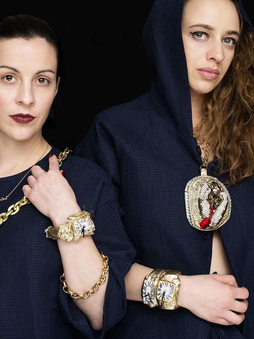 Zwei Rapperin mit dunkelblauen Umhängen und übertrieben großen Goldketten.