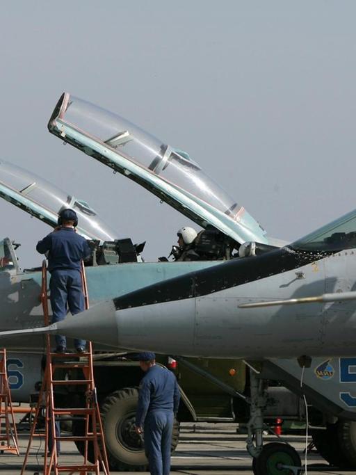 Die Kampfjets werden von Technikern gewartet, die auf roten Leitern an den Cockpits stehen. Darin sitzen Piloten mit Helmen. Die Glasklappen der Cockpits sind geöffnet.