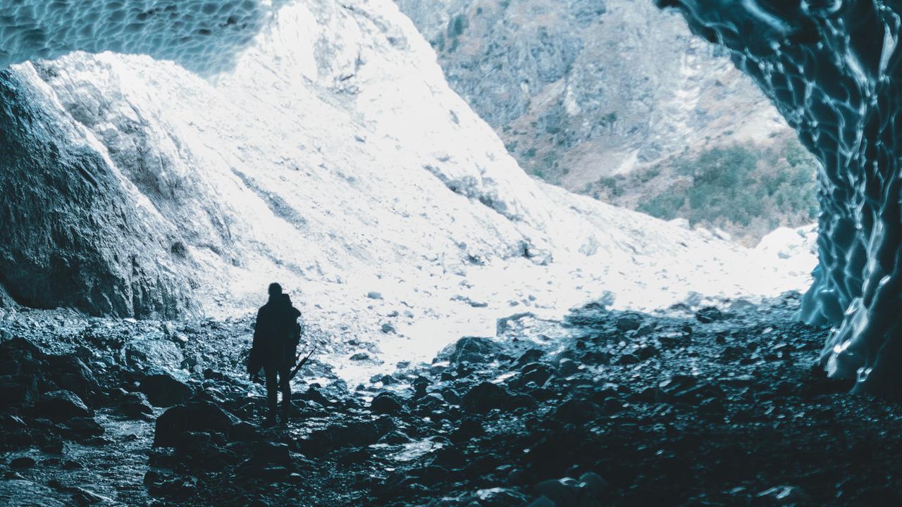 Ein Mann von hinten tritt aus einer Höhle und blickt auf schneebedecktes Gebirge. Ein Mann von hinten tritt aus einer Höhle und blickt auf schneebedecktes Gebirge.