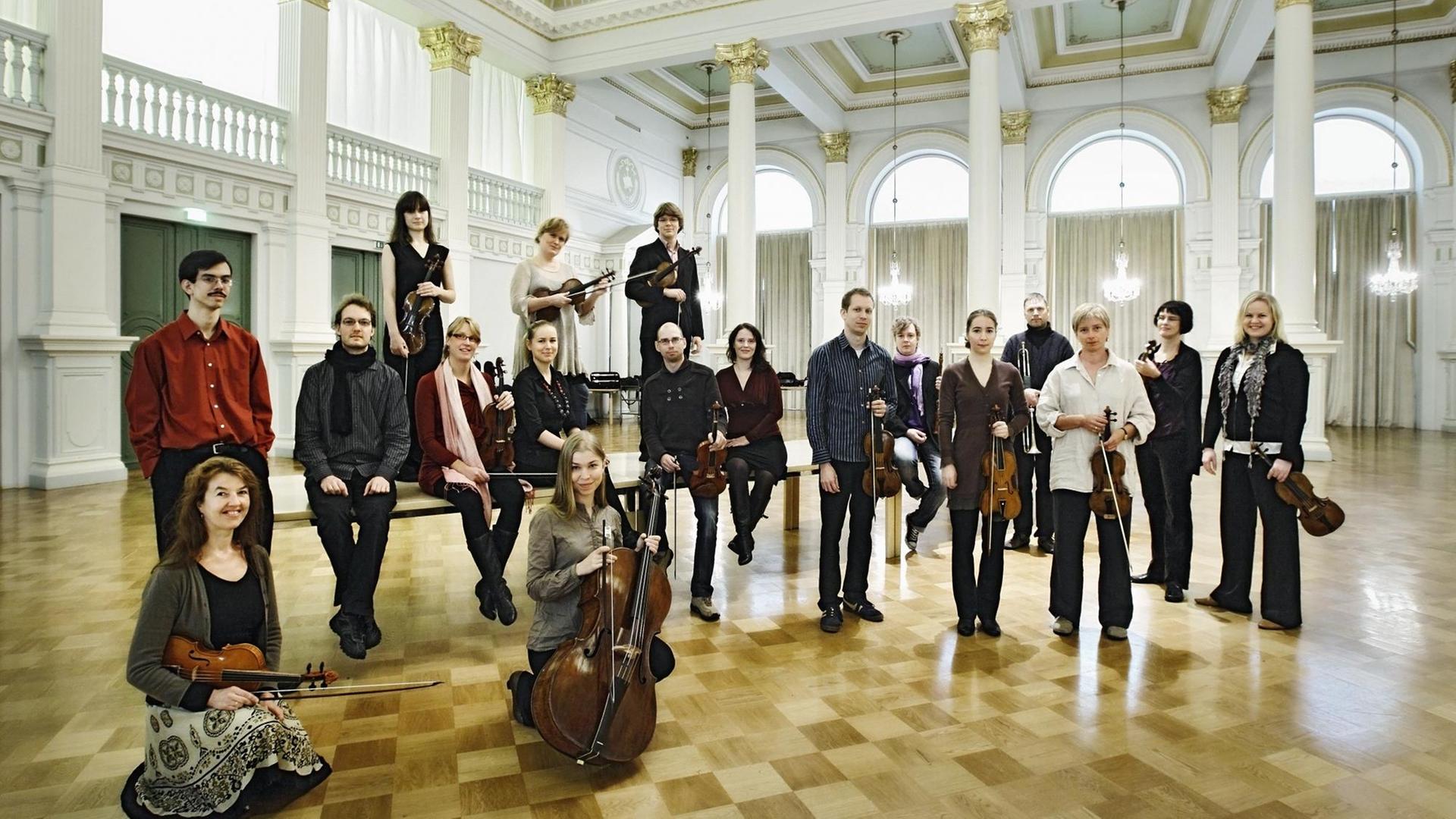 Die Mitglieder des Helsinki Barockorchester sind in einem Halbkreis aufgestellt, manche sitzen auf auf einem Tisch oder knien davor, im Hintergrund ein festlicher Saal mit Säulen und Parkett. Es sind hier 18 Musikerinnen und Musiker in normaler Kleidung, sie halten ihre Instrumente in der Hand. Der Dirigent und Cembalist sitzt links auf einem Tisch inmitten des Ensembles, er trägt ein dunkles, gestreiftes Hemd und einen schwarzen Wollschal um den Hals. Die Mitglieder des Helsinki Barockorchester sind in einem Halbkreis aufgestellt, manche sitzen auf auf einem Tisch oder knien davor, im Hintergrund ein festlicher Saal mit Säulen und Parkett. Es sind hier 18 Musikerinnen und Musiker in normaler Kleidung, sie halten ihre Instrumente in der Hand. Der Dirigent und Cembalist sitzt links auf einem Tisch inmitten des Ensembles, er trägt ein dunkles, gestreiftes Hemd und einen schwarzen Wollschal um den Hals.