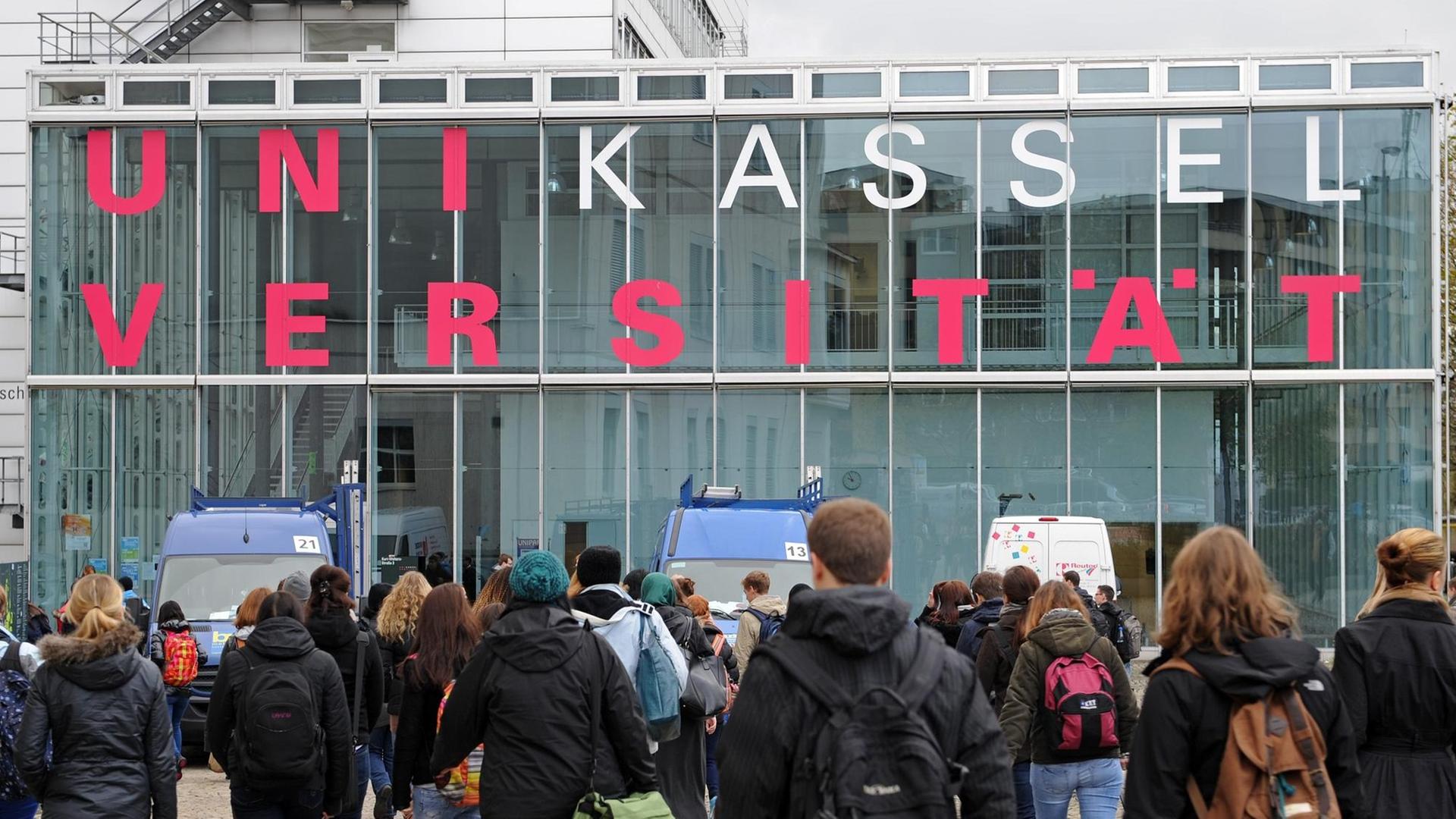 Schriftzug "Universität Kassel" an einem gläsernen Gebäude, davor Studierende, die auf das Gebäude zugehen. Schriftzug "Universität Kassel" an einem gläsernen Gebäude, davor Studierende, die auf das Gebäude zugehen.
