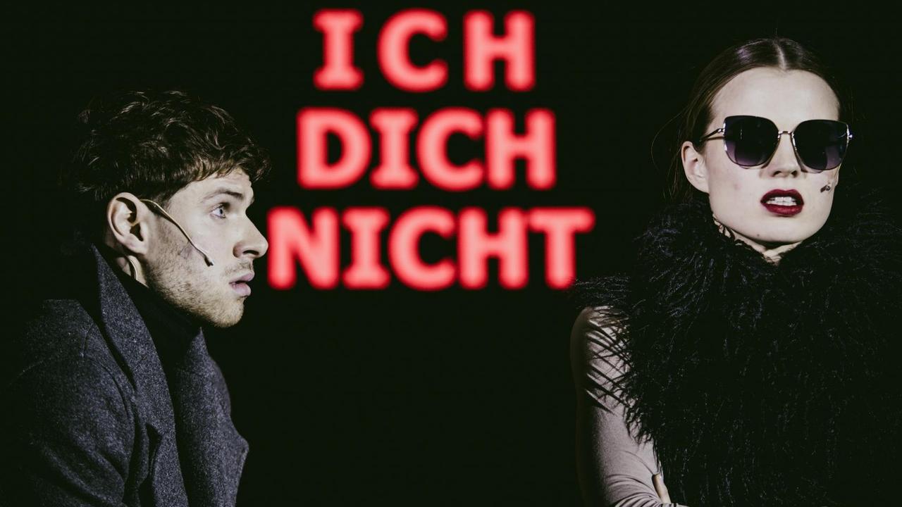 Foto von der Inszenierung: Eine Frau mit Sonnebrille blickt ins Publikum. Ein Mann schaut sie an. Schwarzer Hintergrund. Rote Leuchtbuchstaben: Ich dich nicht. Foto von der Inszenierung: Eine Frau mit Sonnebrille blickt ins Publikum. Ein Mann schaut sie an. Schwarzer Hintergrund. Rote Leuchtbuchstaben: Ich dich nicht.