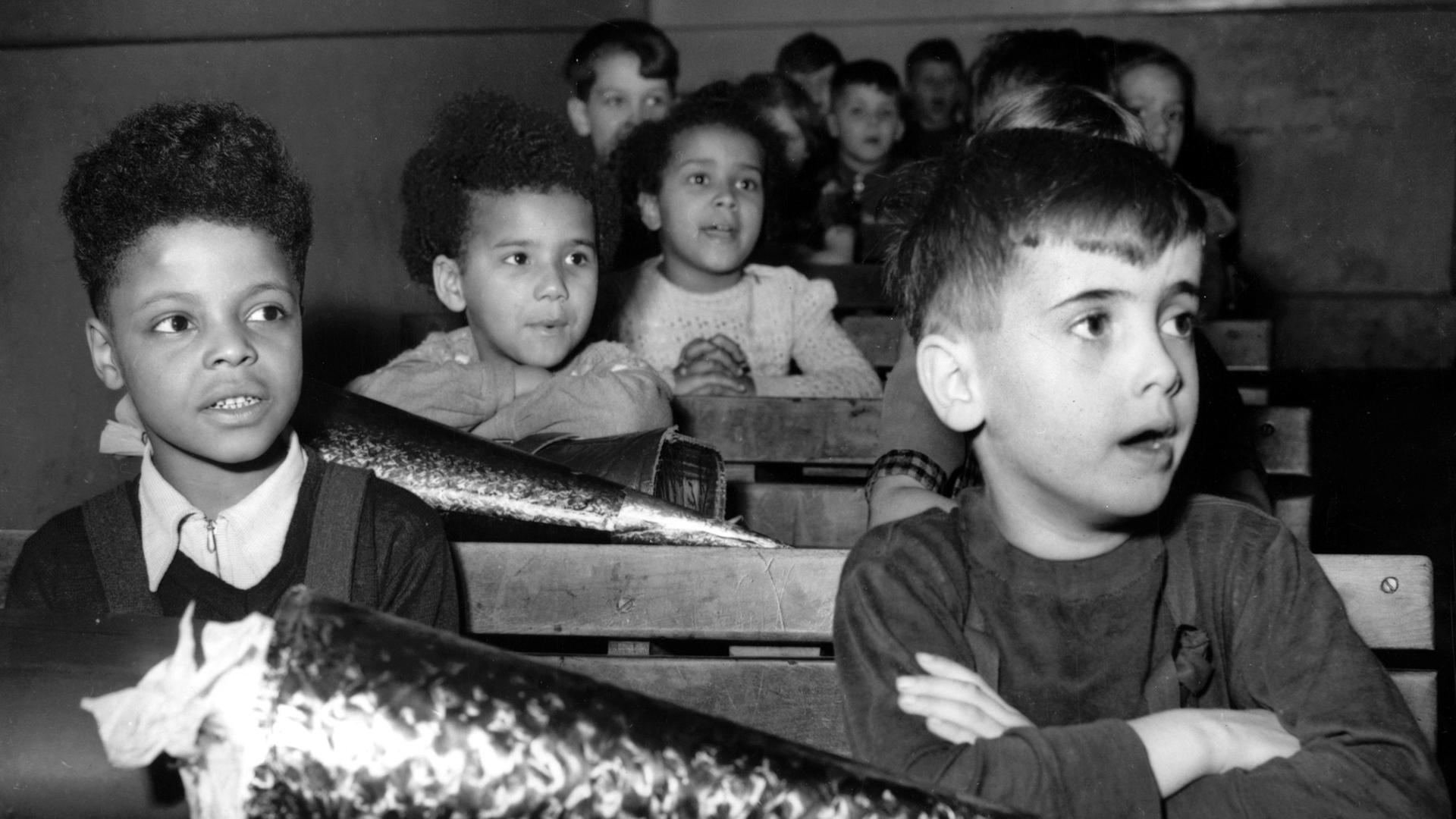 Drei farbige Besatzungskinder und Mitschüler an Tischen im Klassenzimmer, vor ihnen große, glitzernde Schultüten. Drei farbige Besatzungskinder und Mitschüler an Tischen im Klassenzimmer, vor ihnen große, glitzernde Schultüten.
