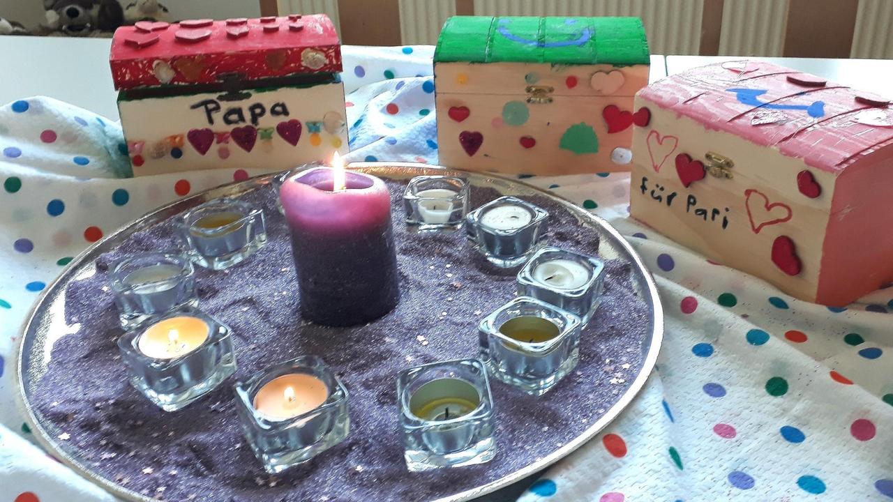 Auf einem Tisch steht ein Tablett mit angezündeten Teelichtern. Dahinter stehen kleine bemalte Holzschachteln auf denen "Papa" und "für Papi" steht. Auf einem Tisch steht ein Tablett mit angezündeten Teelichtern. Dahinter stehen kleine bemalte Holzschachteln auf denen "Papa" und "für Papi" steht.
