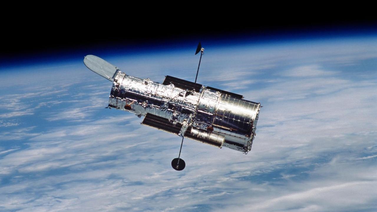 Das Hubble Space Telescope, im Hintergrund die Erde mit Wolken und blauen Schimmern dazwischen, am oberen Bildrand zeigt sich schwarz der Weltraum mit winzigen Sternen.