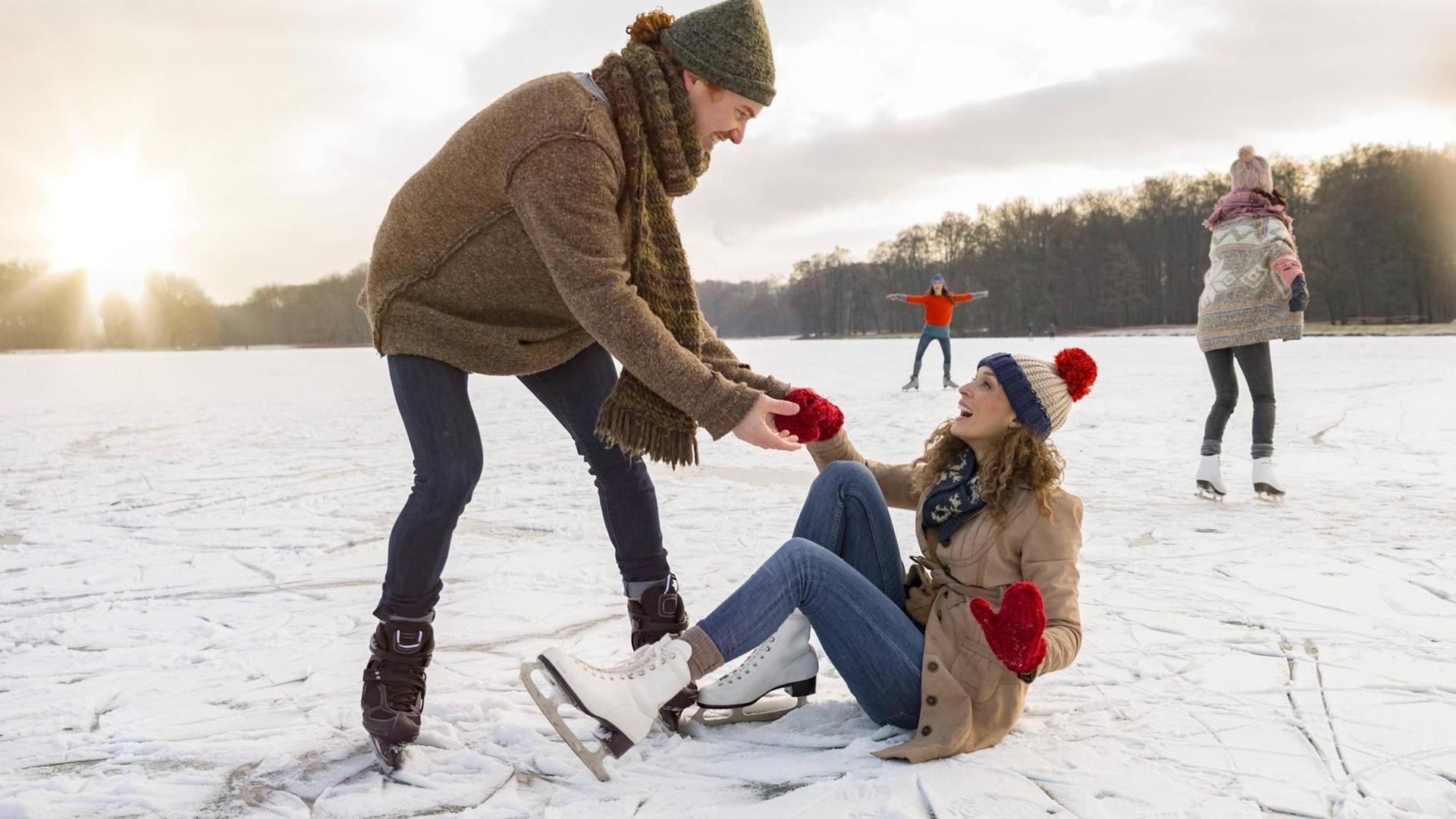 Eine Frau stürzt beim Eislaufen auf denn Boden und ein Mann hilft ihr gerne wieder auf die Beine.