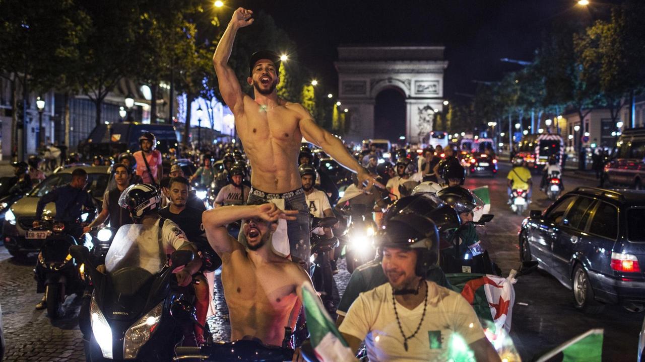 Im Bildzentrum in der Nacht zwei muskulöse Fans mit nacktem Oberkörper auf einem Motorroller, daneben weitere feiernde Kraftradfahrer. Im Hintergrund der Triumphbogen. 