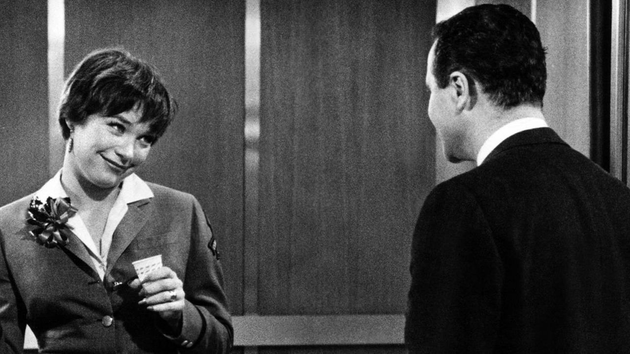 Shirley Maclaine als Fran Kubelik im Film The Apartment USA 1960.  Ein Mann betritt den  Fahrstuhl und Shirley McLain lächelt ihn als Fahrstuhlfhrerin in Uniform freundlich an.
