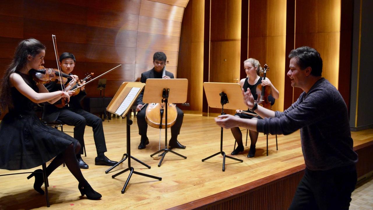 Vier Musiker in Konzertkleidung proben unter Anleitung eines Dozenten auf einer Bühne 