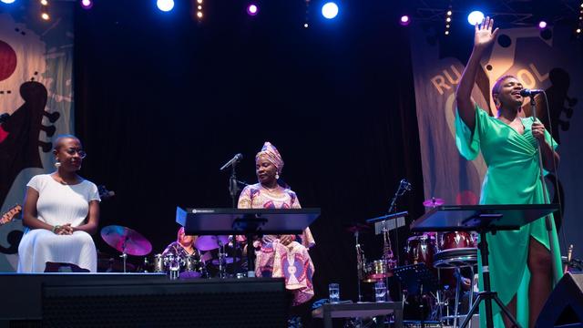 Vier Frauen befinden sich auf einer Bühne. Eine der Frauen steht im grünen Kleid und singt in ein Mikrofon, die rechte Hand dabei zum Ausdruck in die Luft gehoben.