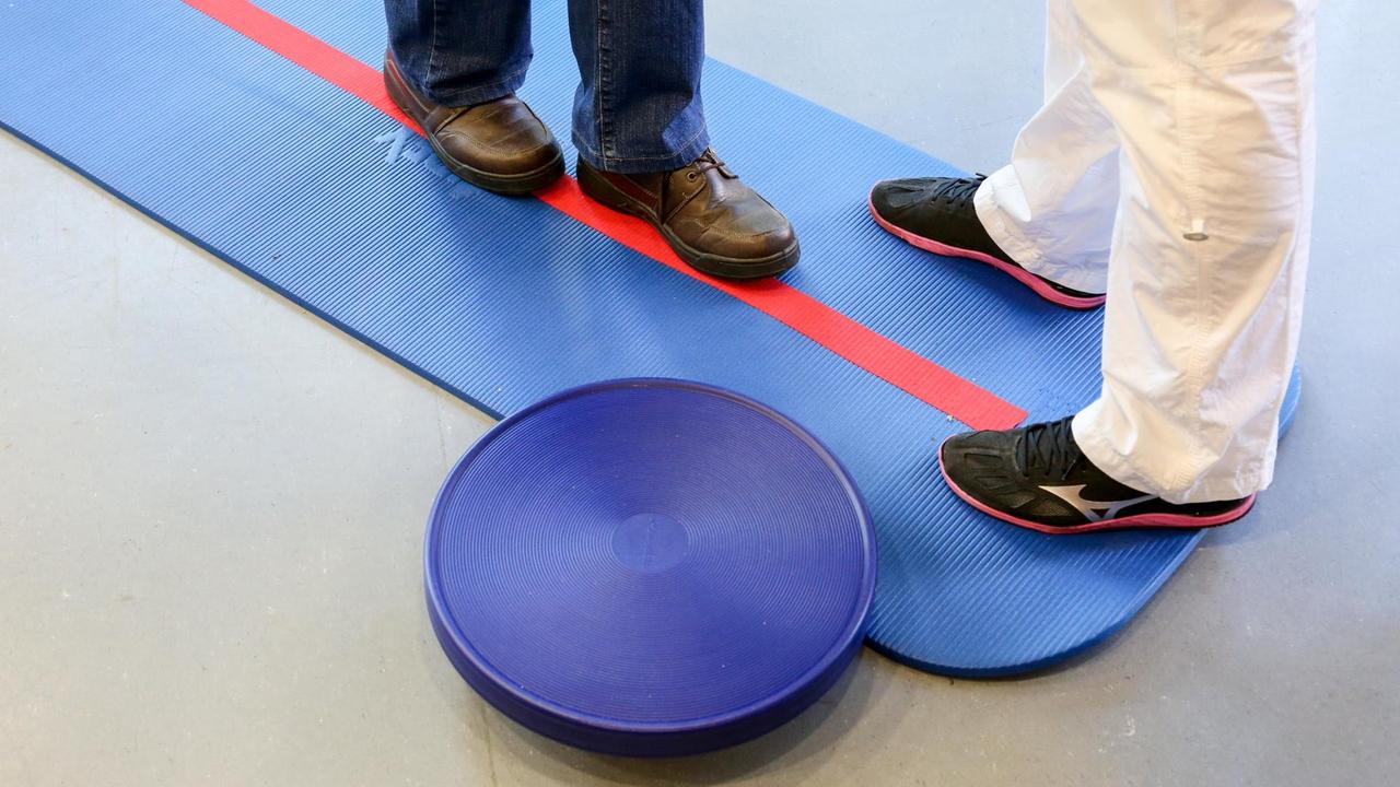 Ein Mensch geht auf einer blauen Matte mit roter Linie, daneben liegt ein Therapiekreisel. Ein Mensch geht auf einer blauen Matte mit roter Linie, daneben liegt ein Therapiekreisel.