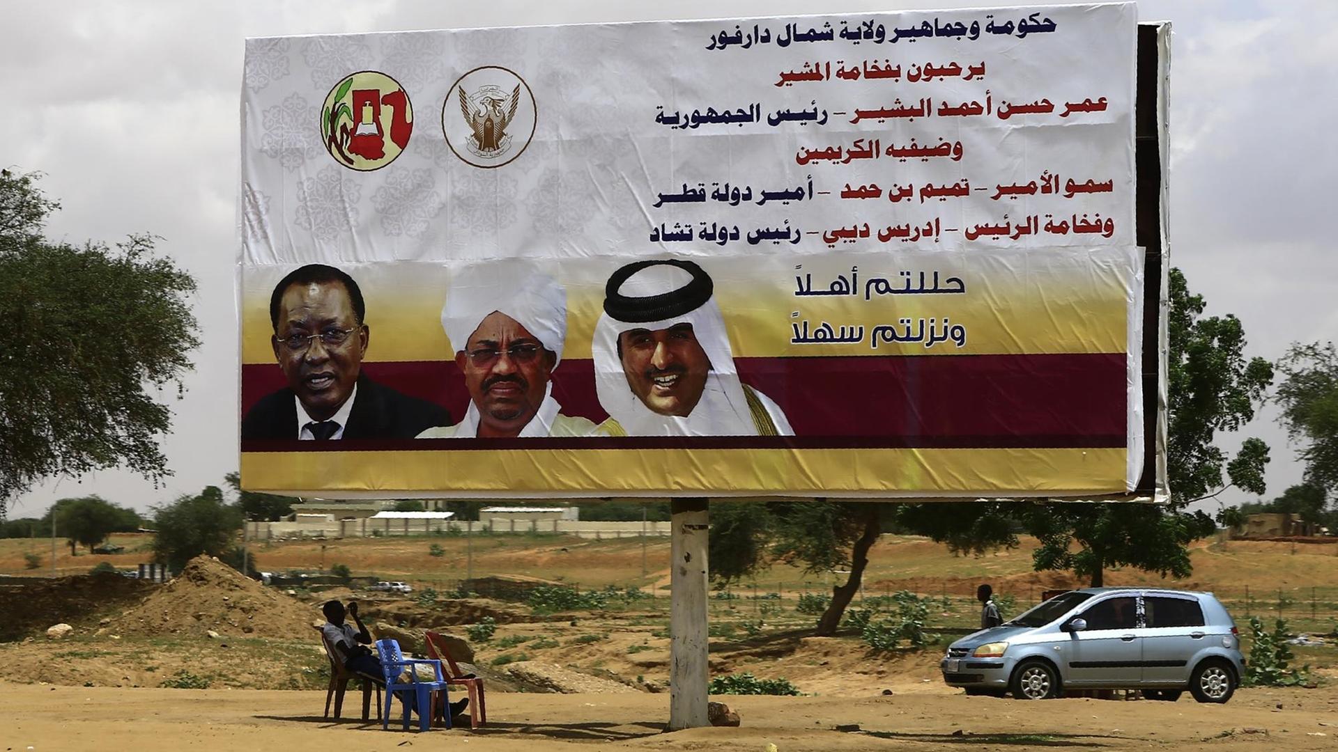 Sudanesische Männer sitzen vor einem Plakat mit den Bildern des sudanesischen Präsidenten Omar al-Bashir (Mitte), dem katarischen Emir Sheikh Tamim bin Hamad al-Thani (r.) und dem Präsidenten des Tschad Idriss Deby (l.) in der El Fasher (Darfur) am 5. September 2016, bevor die politischen Anführer die Gegend besuchen.