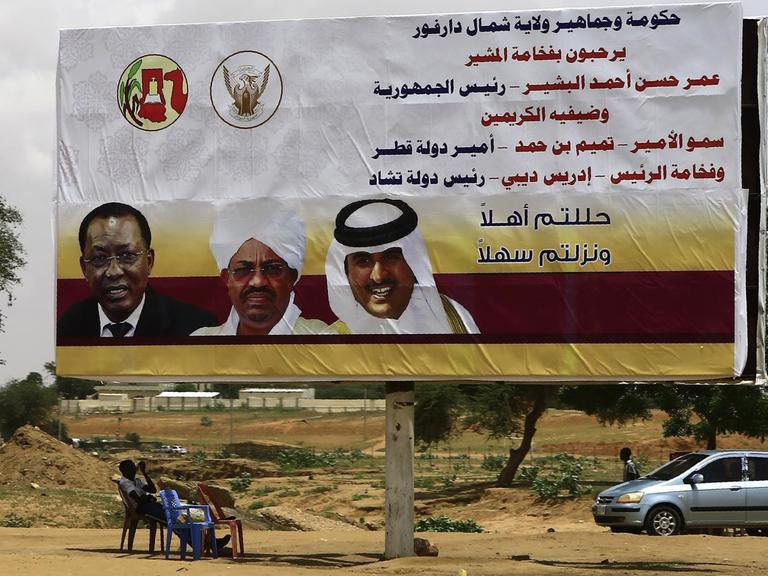 Sudanesische Männer sitzen vor einem Plakat mit den Bildern des sudanesischen Präsidenten Omar al-Bashir (Mitte), dem katarischen Emir Sheikh Tamim bin Hamad al-Thani (r.) und dem Präsidenten des Tschad Idriss Deby (l.) in der El Fasher (Darfur) am 5. September 2016, bevor die politischen Anführer die Gegend besuchen.