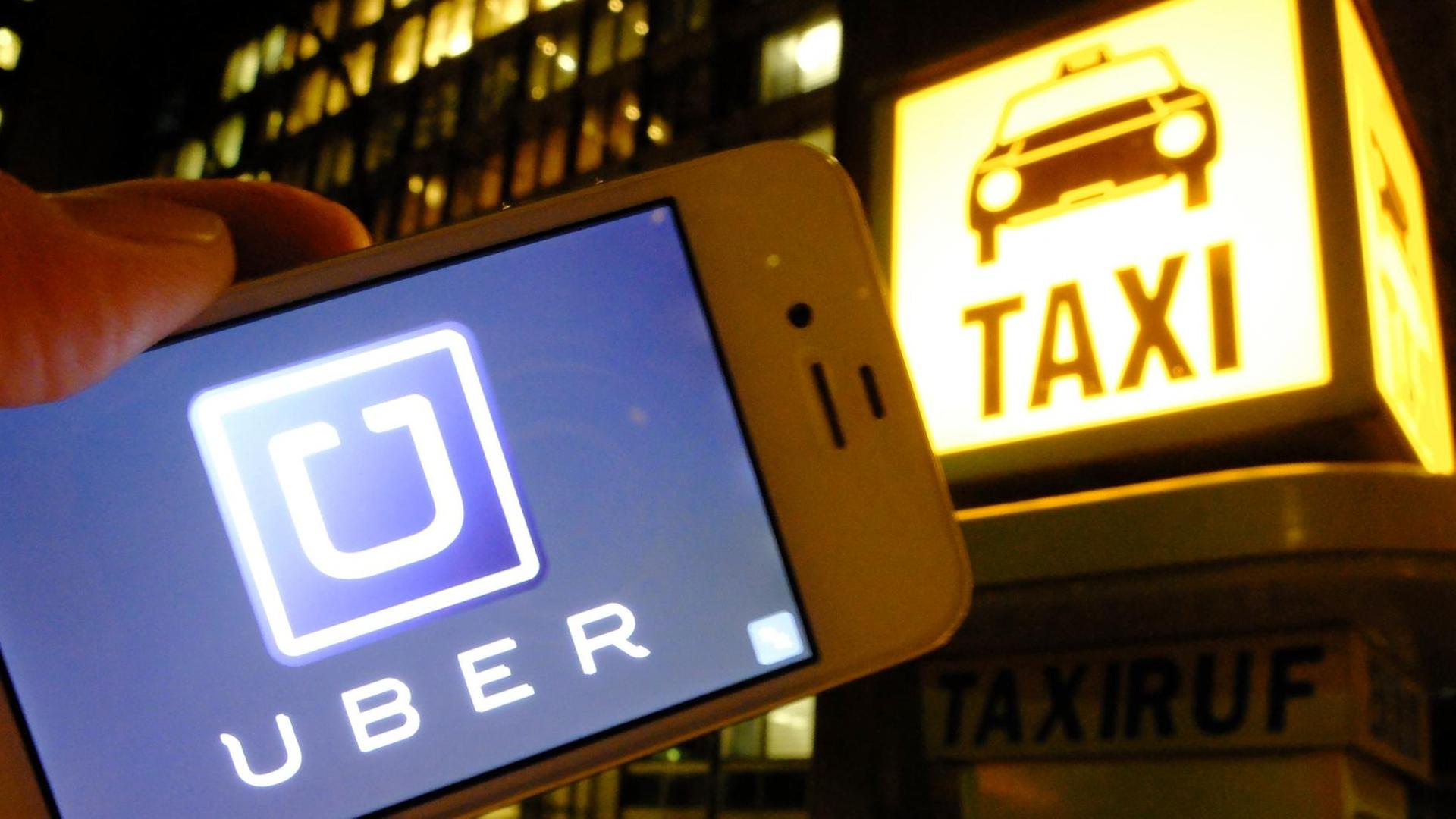 Es ist Nacht in einer Stadt: Eine Hand hält ein Smartphone mit dem Uber-Logo in die Kamera; im Hintergrund ist eine Taxirufsäule mit dem beleuchteten "Taxi"-Logo zu sehen. Es ist Nacht in einer Stadt: Eine Hand hält ein Smartphone mit dem Uber-Logo in die Kamera; im Hintergrund ist eine Taxirufsäule mit dem beleuchteten "Taxi"-Logo zu sehen.