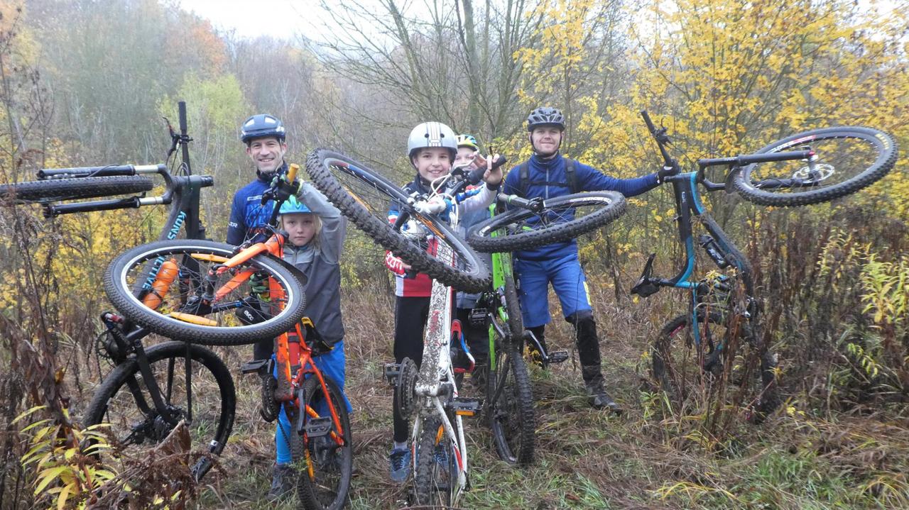Eine Gruppe von Kindern und einem Trainer ist mit ihren Mountainbikes im Wald zu sehen. 