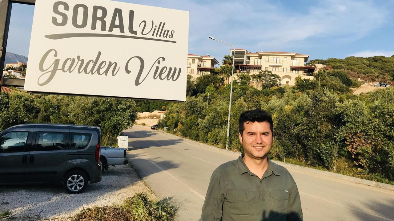 Vor einer grünen sonnenbeschienenen hügeligen Landschaft mit prächtigen Villen steht am Rand einer Straße ein junger schwarzhaariger Mann mit olivgrünem Hemd und lächelt. Hinter ihm steht ein großes Straßenschild in Weíß auf dem steht: "Soral Villa. Garden View". Vor einer grünen sonnenbeschienenen hügeligen Landschaft mit prächtigen Villen steht am Rand einer Straße ein junger schwarzhaariger Mann mit olivgrünem Hemd und lächelt. Hinter ihm steht ein großes Straßenschild in Weíß auf dem steht: "Soral Villa. Garden View".