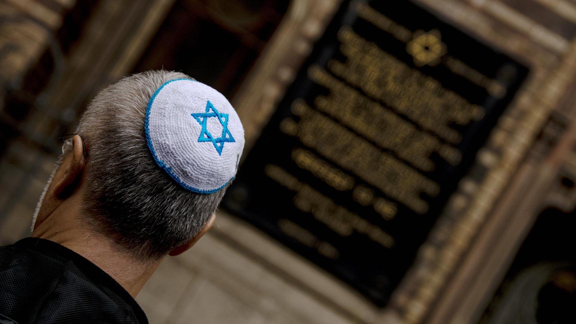 Rückansicht eines jüdischen Mannes mit Kippa, auf der sich ein blauer Judenstern auf weißem Grund befindet. Im Hintergrund zu sehen ist eine Fassadeninschrift. Rückansicht eines jüdischen Mannes mit Kippa, auf der sich ein blauer Judenstern auf weißem Grund befindet. Im Hintergrund zu sehen ist eine Fassadeninschrift.