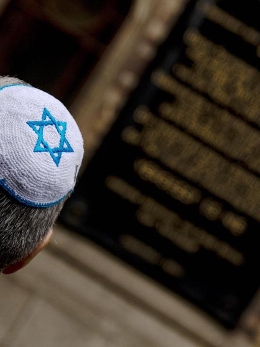 Rückansicht eines jüdischen Mannes mit Kippa, auf der sich ein blauer Judenstern auf weißem Grund befindet. Im Hintergrund zu sehen ist eine Fassadeninschrift.