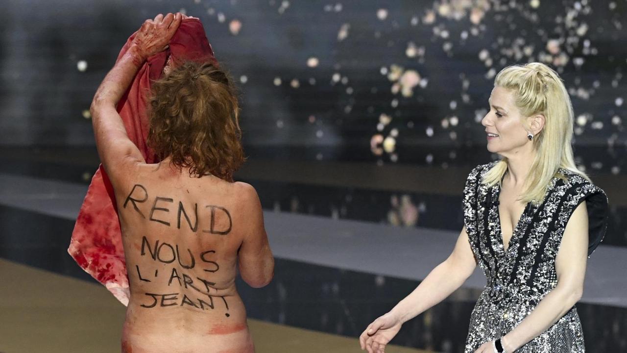 Die Schauspielerin Corinne Masiero steht nackt mit dem Rücken zur Kamera auf der Bühne bei der Verleihung des César. Auf Französisch steht auf ihrem Rücken "Gib uns die Kunst zurück, Jean!" Rechts von ihr steht angezogen die Moderatorin.