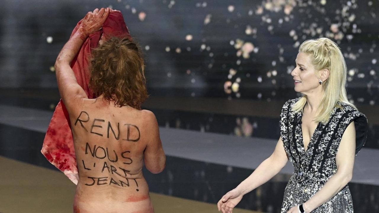 Die Schauspielerin Corinne Masiero steht nackt mit dem Rücken zur Kamera auf der Bühne bei der Verleihung des César. Auf Französisch steht auf ihrem Rücken "Gib uns die Kunst zurück, Jean!" Rechts von ihr steht angezogen die Moderatorin. Die Schauspielerin Corinne Masiero steht nackt mit dem Rücken zur Kamera auf der Bühne bei der Verleihung des César. Auf Französisch steht auf ihrem Rücken "Gib uns die Kunst zurück, Jean!" Rechts von ihr steht angezogen die Moderatorin.