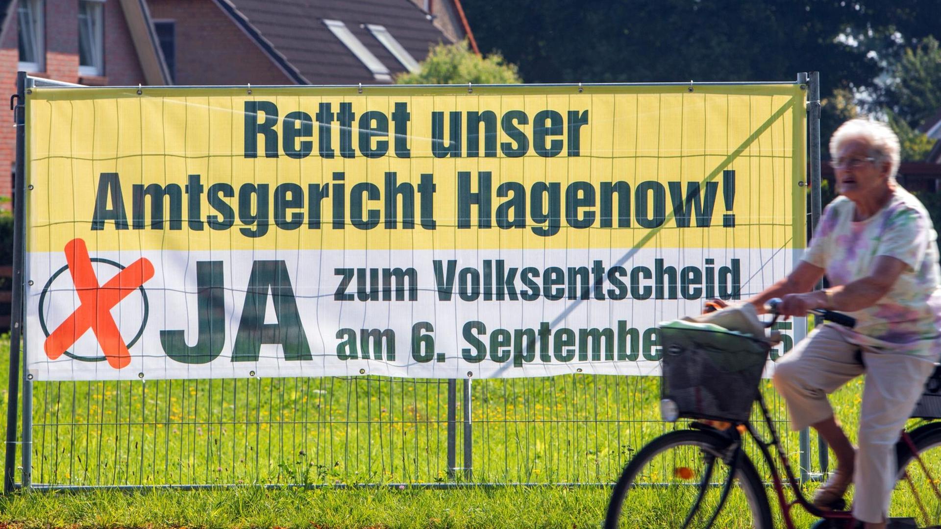Plakat hängt an einem Zaun, eine Frau auf einem Rad fährt vorbei, Aufschrift auf dem Plakat: "Rettet unser Amtsgericht Hagenow! Ja zum Volksentscheid am 6. September"